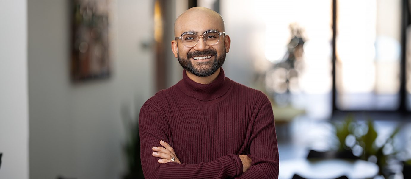 A person wearing glasses and a maroon sweater smiles with arms crossed, standing confidently indoors. They seem like they could easily guide you through any main navigation setting, their warmth radiating in the blurred background.