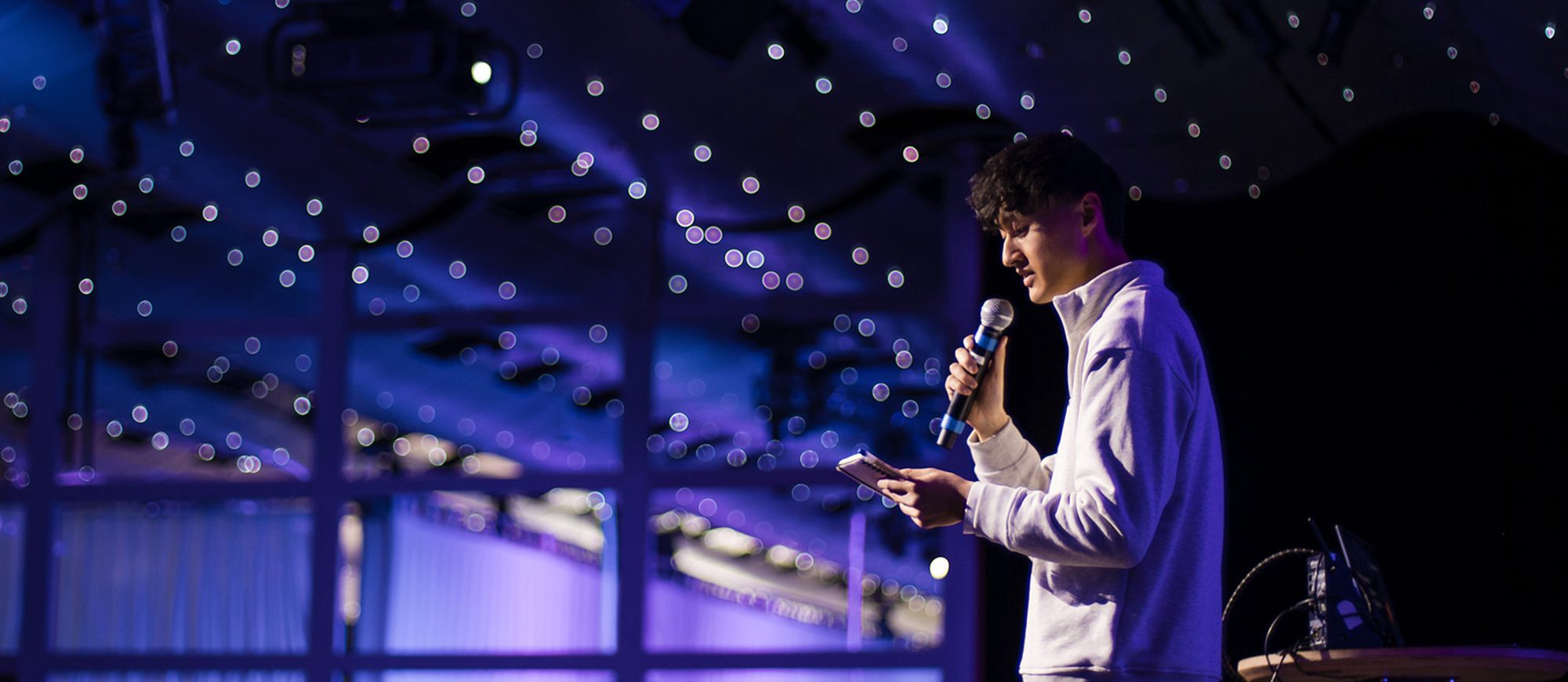 A person in a white jacket speaks confidently into a microphone on stage, expertly guiding the audience through the main navigation of the evening's event, all set against a mesmerizing purple-lit background.