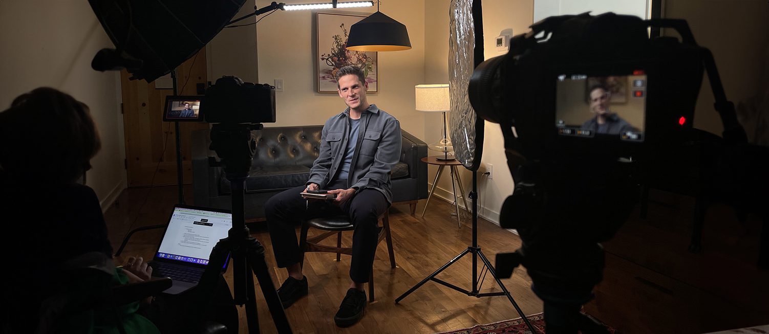 A person sits on a couch holding a tablet in a dimly lit room, navigating through the website interface, while facing a camera setup complete with lights and a laptop.