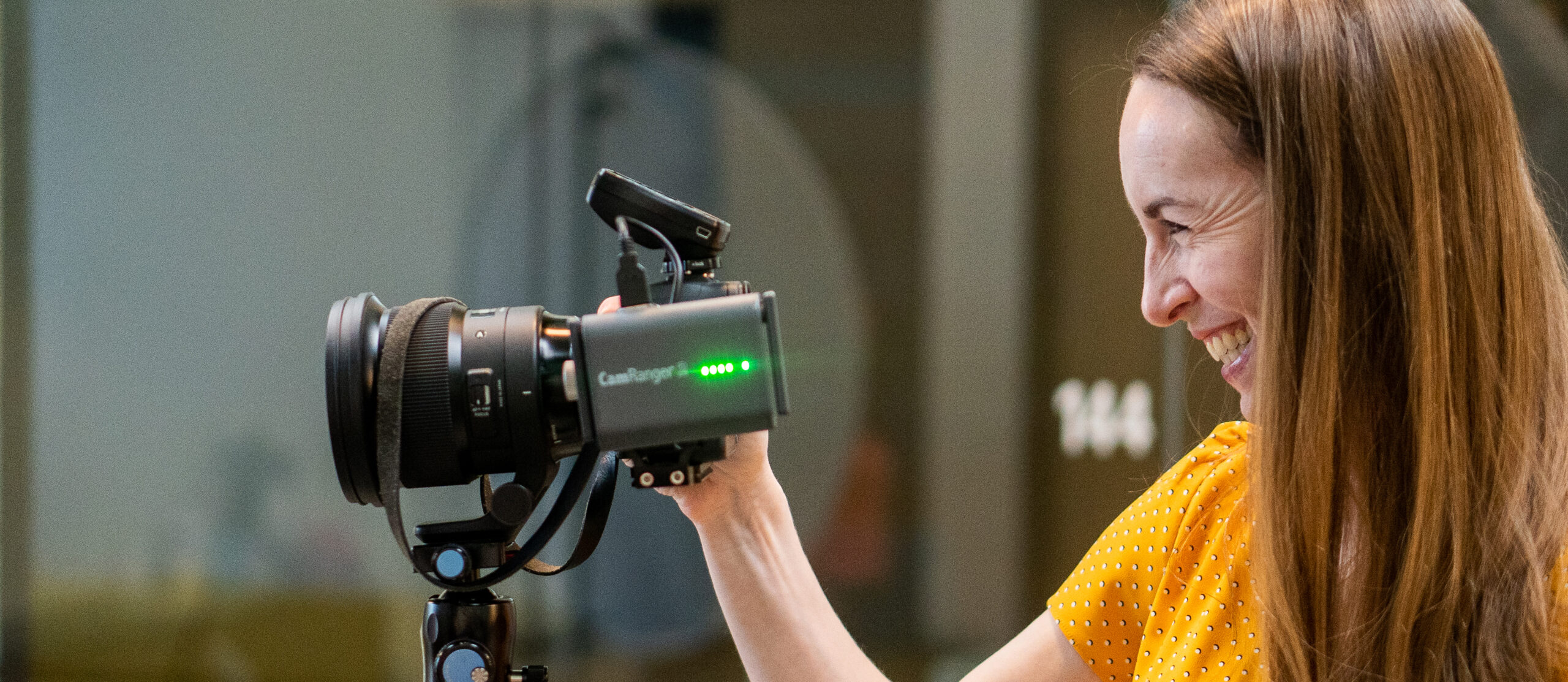 A woman in a yellow polka dot shirt smiles and looks through a professional camera mounted on a tripod indoors, capturing moments with skillful precision.