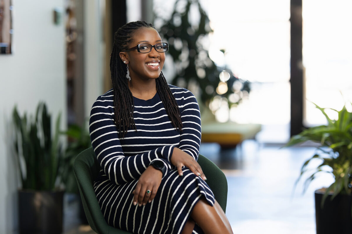 A smiling woman with glasses, wearing a striped dress, sitting in a green chair in a modern office space for her Denver headshots.