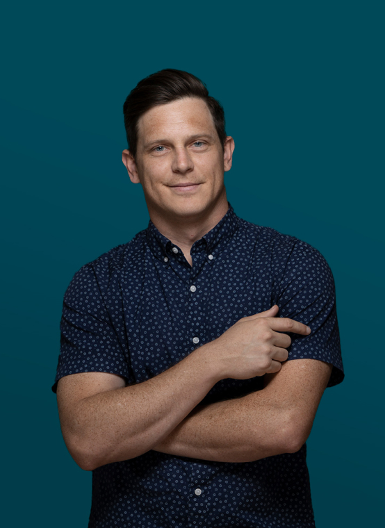 A man with short dark hair and a blue patterned shirt stands against a teal background, crossing one arm and pointing with the other. This striking image by Denver Headshot Co captures confidence and style effortlessly.