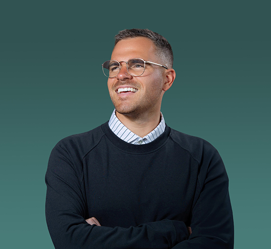 A person with short hair and glasses is smiling while looking slightly to the side. They are wearing a dark sweater over a collared shirt against a teal background, showcasing the professional touch of Denver Headshot Co.