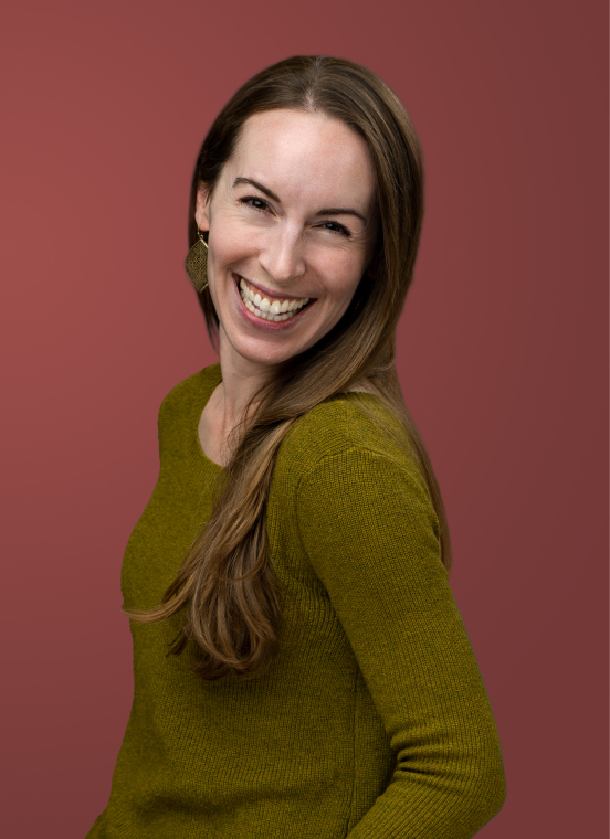 A woman with long brown hair wearing a green sweater smiles and looks sideways against a red background, captured brilliantly by Denver Headshot Co.