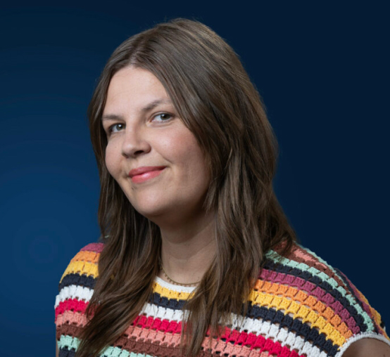 A woman with long brown hair is smiling subtly. She is wearing a colorful striped knit top and is posed against a dark blue background, captured perfectly by Denver Headshot Co.