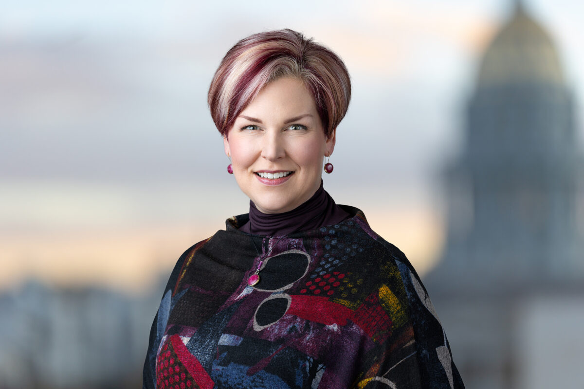 A woman with short, highlighted hair wearing a colorful patterned top smiles with a blurred domed building in the background, capturing the spirit of a Denver Headshot Co session.