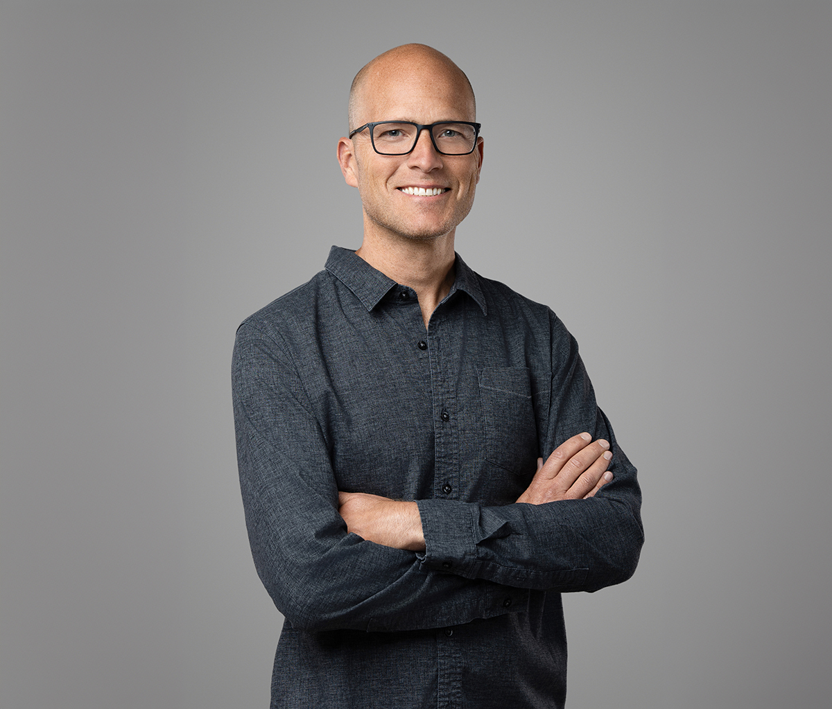 A bald man wearing glasses and a dark button-up shirt stands smiling with arms crossed against a plain gray background, perfectly capturing the professionalism championed by Denver Headshot Co.