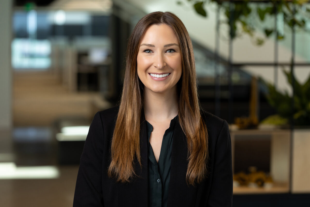 A woman with long brown hair wearing a black blazer smiles in a modern office environment, captured flawlessly by Denver Headshot Co.