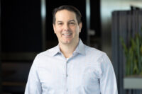 A person with short dark hair is standing indoors wearing a light-colored, button-down shirt, smiling at the camera—a classic Denver Headshot Co portrait.