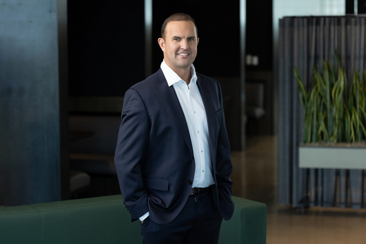 A man in a business suit stands indoors near a green couch and a plant, smiling and looking at the camera, showcasing the professional touch of Denver Headshot Co.