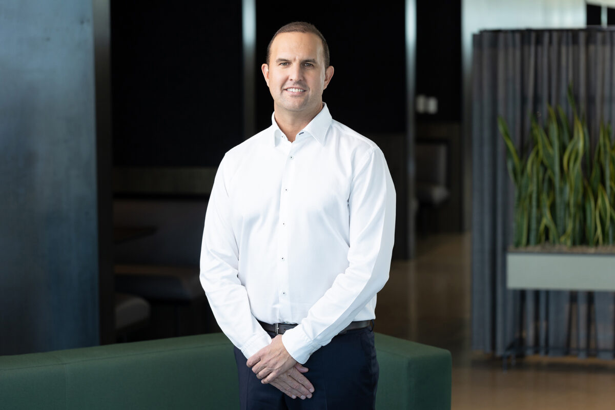 A man in a white shirt stands indoors, smiling at the camera with hands clasped in front. The background includes office furniture and a plant, perfectly captured by Denver Headshot Co.