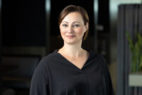 A woman with short brown hair and a black shirt smiles slightly while standing indoors with a blurred background, captured in a professional shot by Denver Headshot Co.