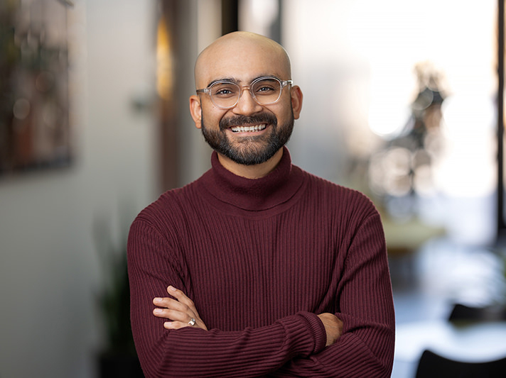 A bald man with glasses and a neat beard, wearing a maroon sweater, stands indoors with crossed arms, smiling at the camera in a professional Denver Headshot Co setting.