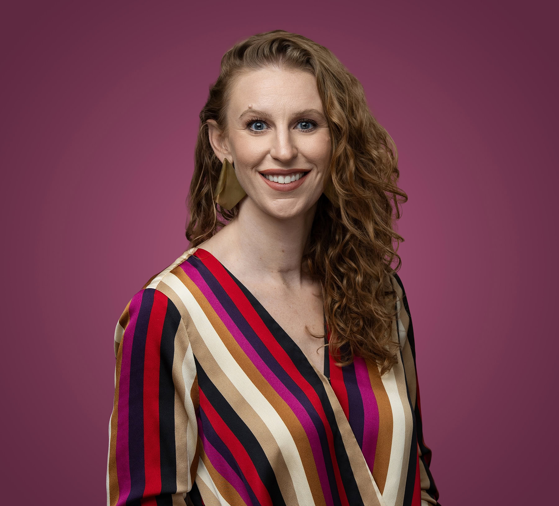 A person with long, curly hair wearing a striped, colorful shirt stands smiling against a deep magenta background, perfectly capturing the vibrant essence of a Denver Headshot Co photo shoot.