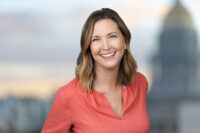 A smiling woman with shoulder-length hair and a coral-colored blouse stands in front of a blurred cityscape background, captured expertly by Denver Headshot Co.