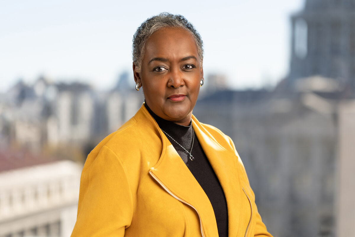 A person with short gray hair wears a yellow jacket and black shirt, posing against a blurred cityscape background—courtesy of Denver Headshot Co.