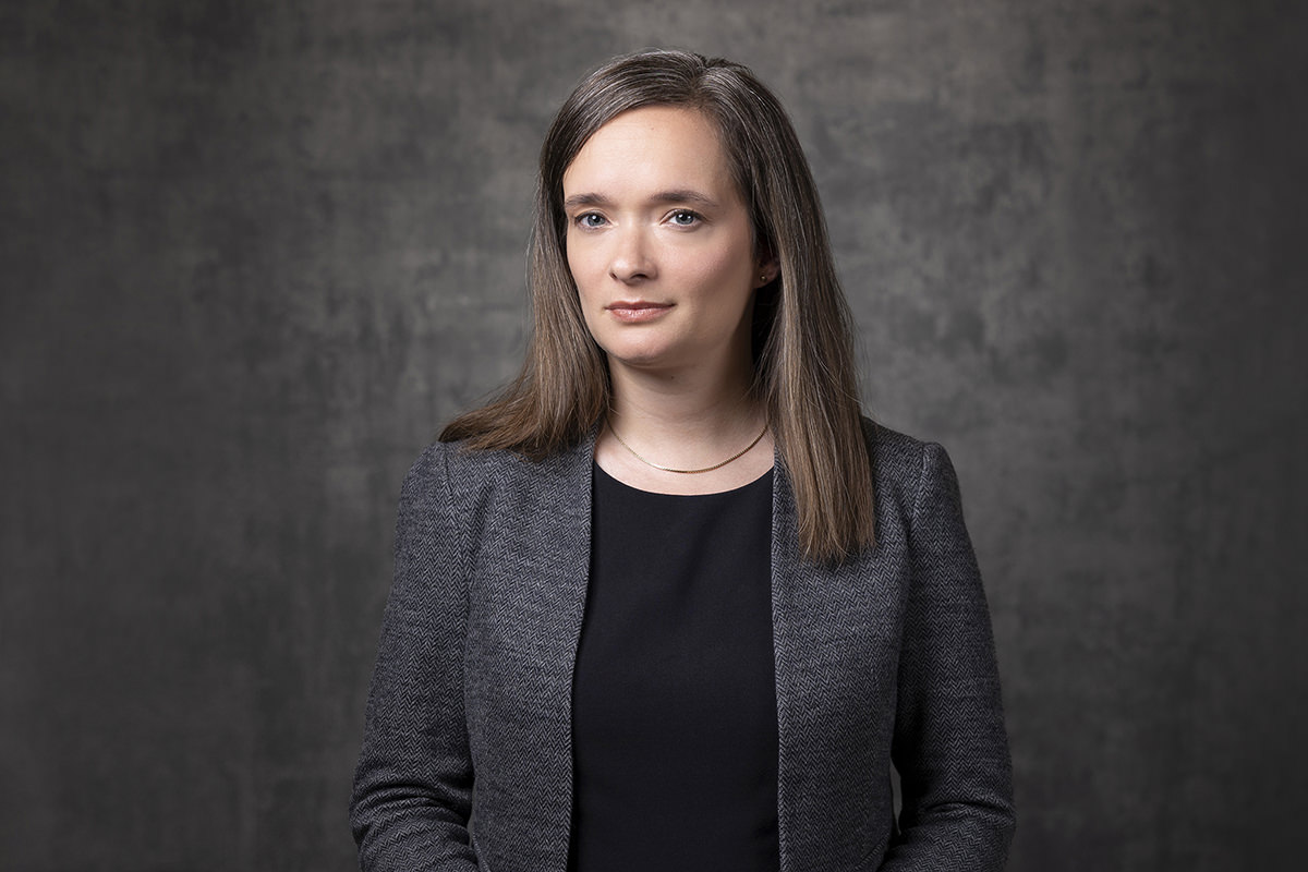 A woman with long, straight hair wearing a dark gray blazer and black top stands against a dark, textured background, looking directly at the camera. This striking image captures the quality of a Denver Headshot Co portrait.