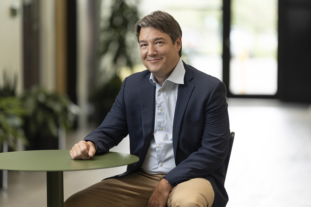 A man wearing a suit jacket and light blue shirt sits at a round green table in a bright room with glass doors in the background, captured perfectly by Denver Headshot Co.