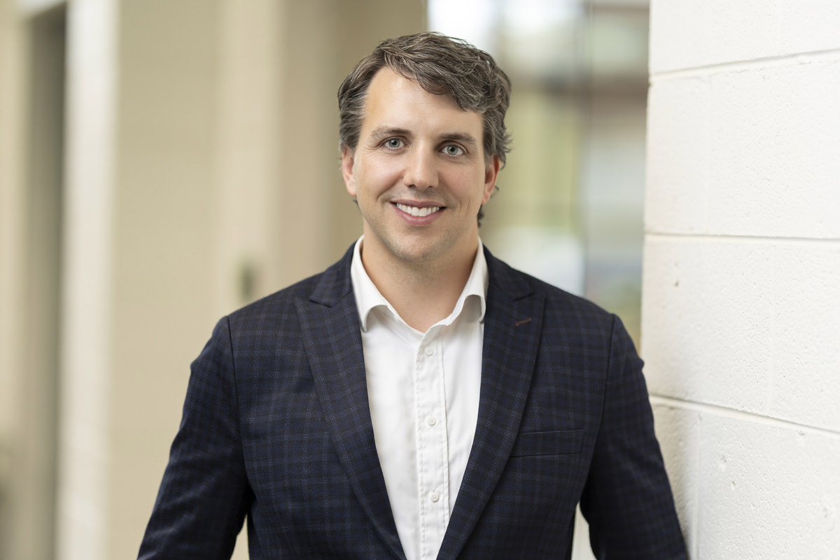 A person with short, wavy hair, dressed in a dark plaid blazer and white shirt, stands indoors near a white brick wall, smiling at the camera—a perfect moment captured by Denver Headshot Co.