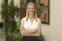 A woman with long blonde hair and a white sleeveless blouse stands indoors with her arms crossed, smiling at the camera. Behind her, plants and a colorful wall hanging add charm to the scene captured perfectly by Denver Headshot Co.