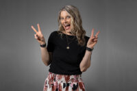 A person with long, wavy hair wearing a black shirt and patterned pants smiles broadly, holding up two peace signs with both hands against a gray background in a Denver Headshot Co session.