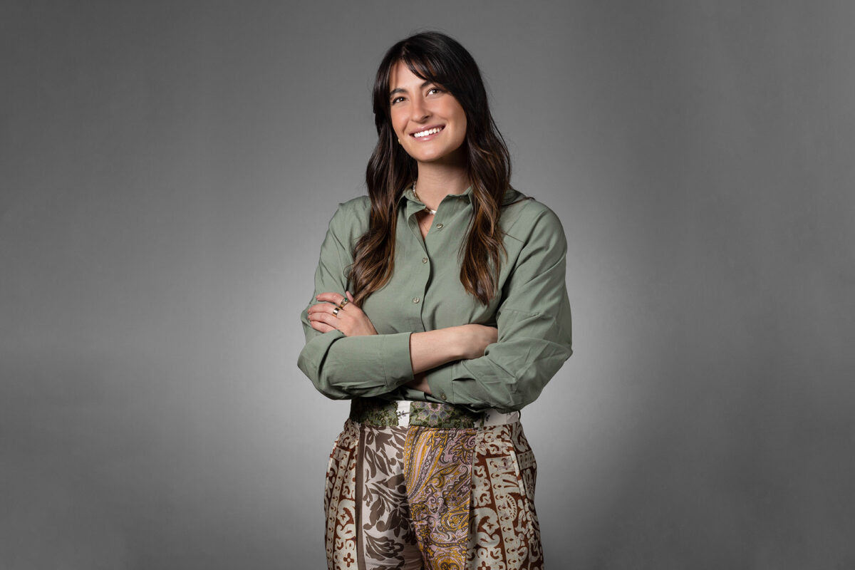 A person with long, dark hair wearing a green shirt and patterned pants stands against a gray background, smiling with arms crossed—a perfect capture from Denver Headshot Co.