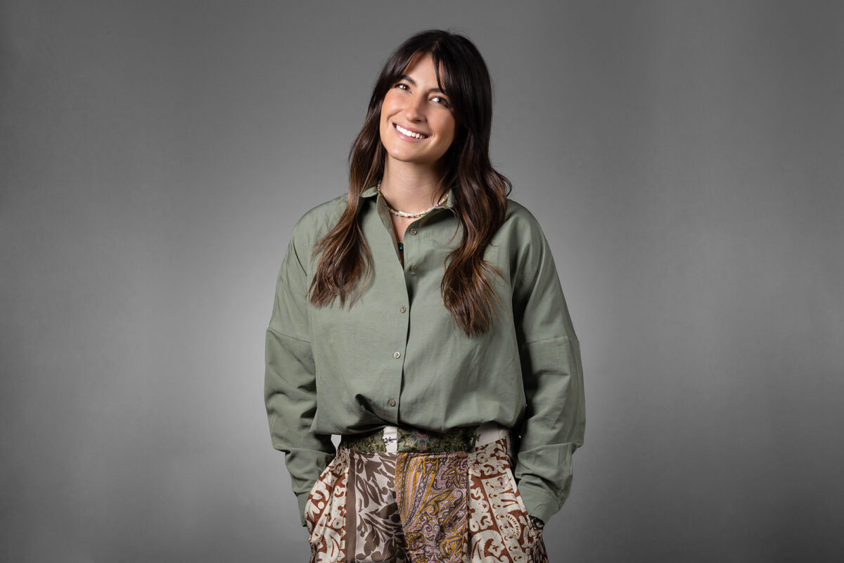 A person with long, dark hair wearing a green shirt and patterned pants stands against a gray background, smiling with hands in pockets—an expert work by Denver Headshot Co.