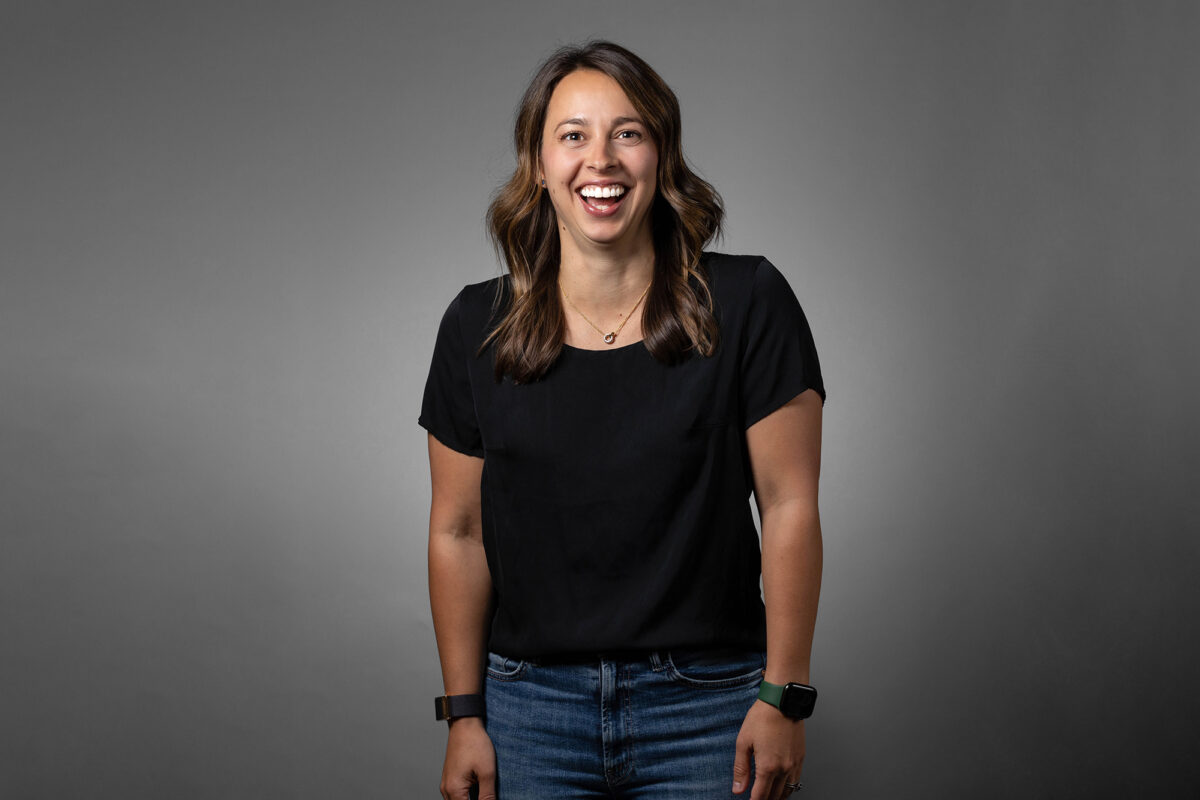 A person stands against a grey background, smiling while wearing a black shirt, blue jeans, and two wristbands. This portrait perfectly captures the professionalism Denver Headshot Co is known for.