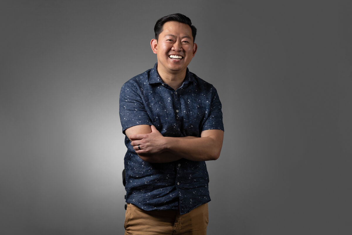 A person wearing a navy blue shirt and tan pants stands with arms crossed, smiling confidently in front of a grey background, photographed by Denver Headshot Co.