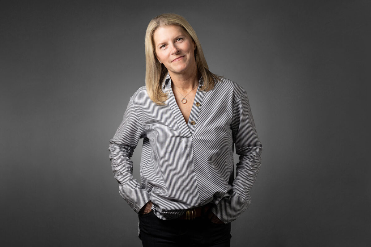 A person with long blonde hair, wearing a gray striped shirt and black pants, poses confidently with hands in pockets against a plain gray background—a perfect example of professional photography by Denver Headshot Co.