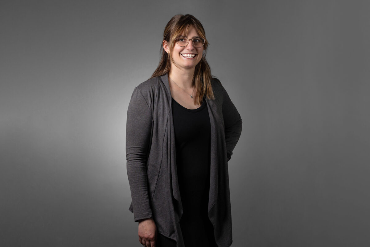 A woman with long hair, glasses, and wearing a dark outfit and gray cardigan stands against a plain gray background, smiling with one hand on her hip. Captured by Denver Headshot Co, the image perfectly encapsulates her professional yet approachable demeanor.