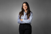 A person with long dark hair, dressed in a light blue striped shirt and black pants, stands against a gray background with their arms crossed, smiling—captured perfectly by Denver Headshot Co.