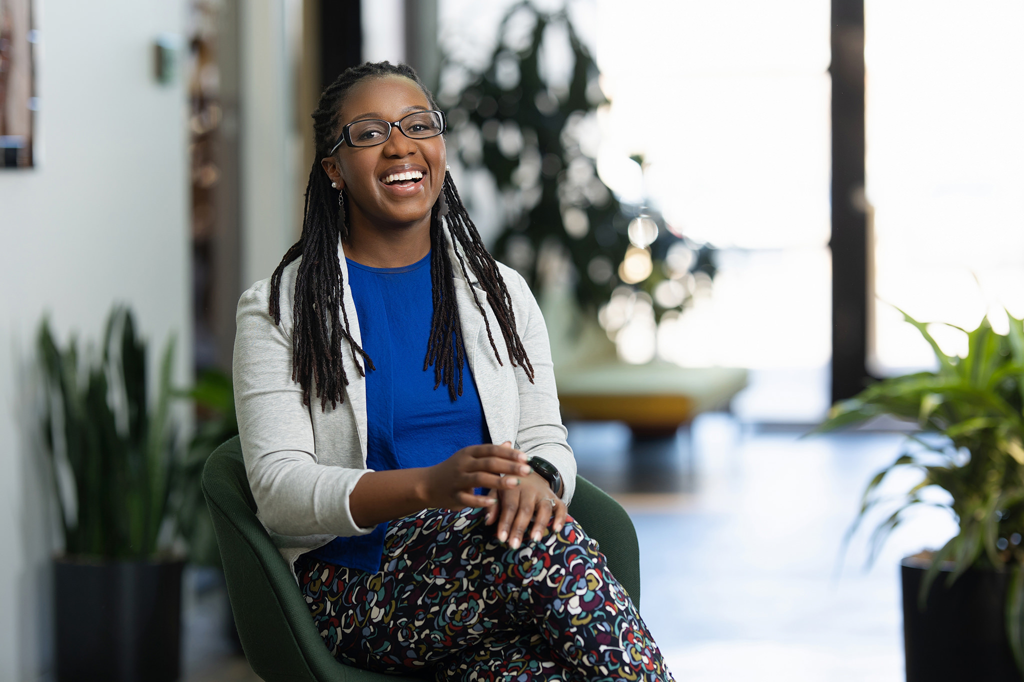 A woman wearing glasses, a light-colored jacket, and patterned pants sits on a green chair, smiling in a brightly lit room with plants in the background—a perfect scene captured by Denver Headshot Co.