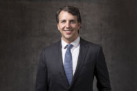 A man in a business suit and tie, expertly captured by Denver Headshot Co, stands against a dark, textured background, smiling at the camera.