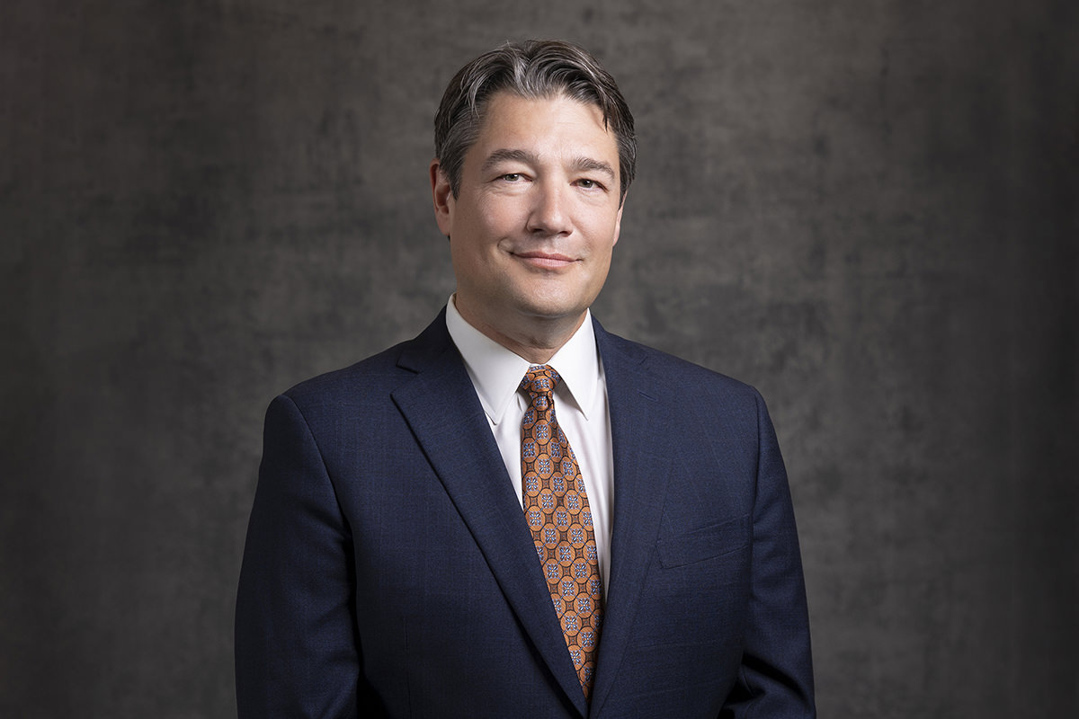 A person wearing a dark suit, white shirt, and patterned tie stands in front of a gray background, facing the camera with a neutral expression. This professional portrait was expertly captured by Denver Headshot Co.