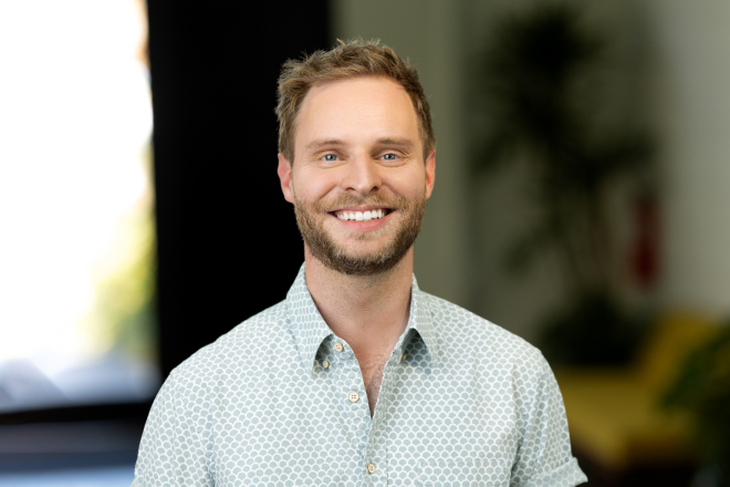 A smiling man with short hair and a beard wears a patterned shirt and stands indoors, captured in a sharp image by Denver Headshot Co., with a beautifully blurred background.