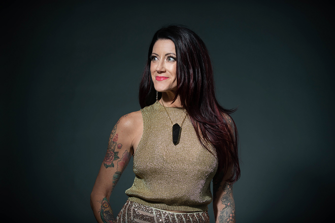 A woman with long dark hair and tattoos wears a metallic sleeveless top, standing against a dark background—a striking image captured by Denver Headshot Co.