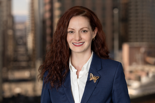 A person with long red hair wearing a blue blazer and a butterfly brooch smiles in front of a blurred cityscape background, capturing the professional touch of Denver Headshot Co.