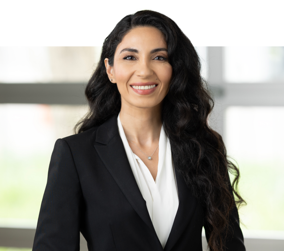 A woman with long, wavy dark hair is standing and smiling. She is wearing a black blazer and white blouse. The background is blurred, capturing the professionalism that Denver Headshot Co excels at.