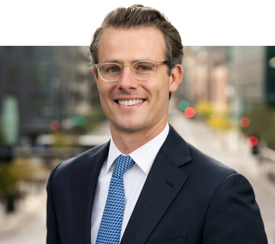 A man wearing glasses, a dark suit, white shirt, and blue tie stands smiling in a cityscape background—a perfect snapshot crafted by Denver Headshot Co.