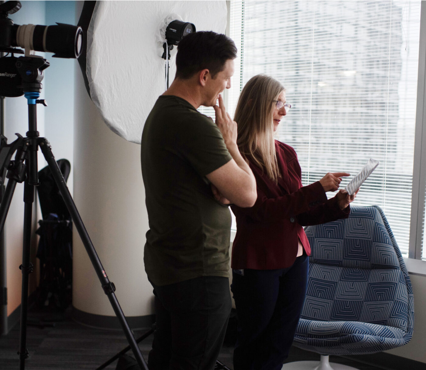 A man and a woman are standing next to a large photography light, engaged in conversation at the Denver Headshot Co's modern office. The woman is holding and pointing to a document, illustrating their discussion.