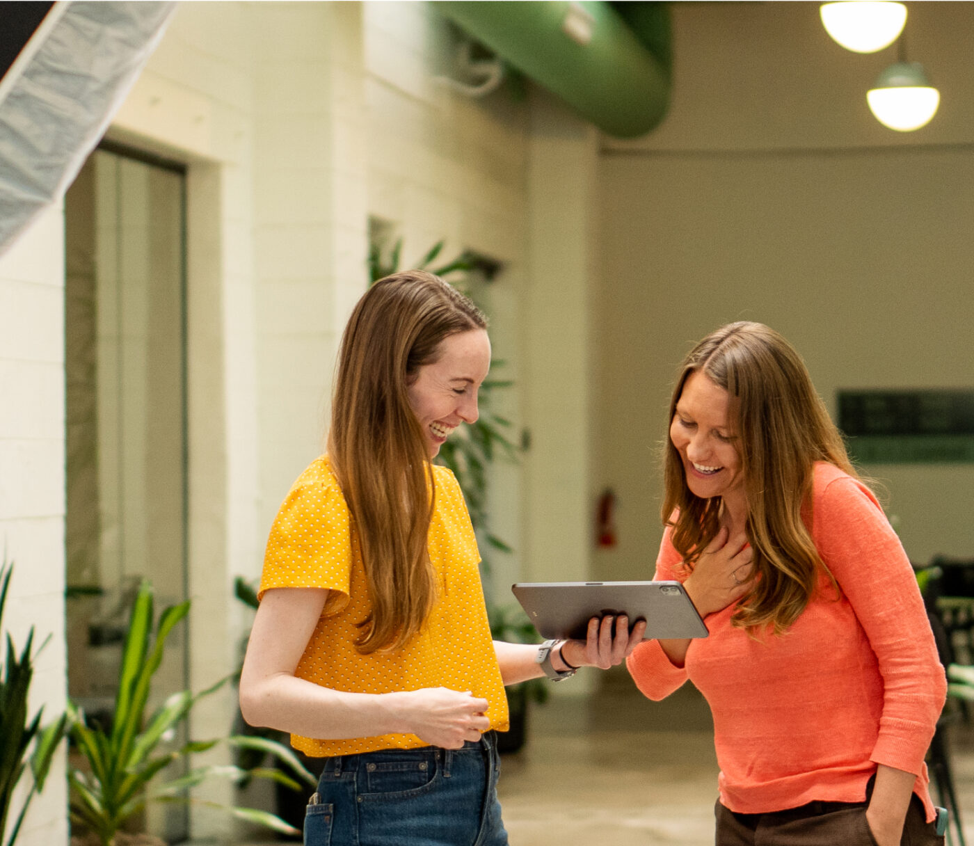 Two women from Denver Headshot Co are smiling and looking at a tablet in a brightly lit indoor space. One woman is wearing a yellow shirt, and the other is wearing an orange shirt.