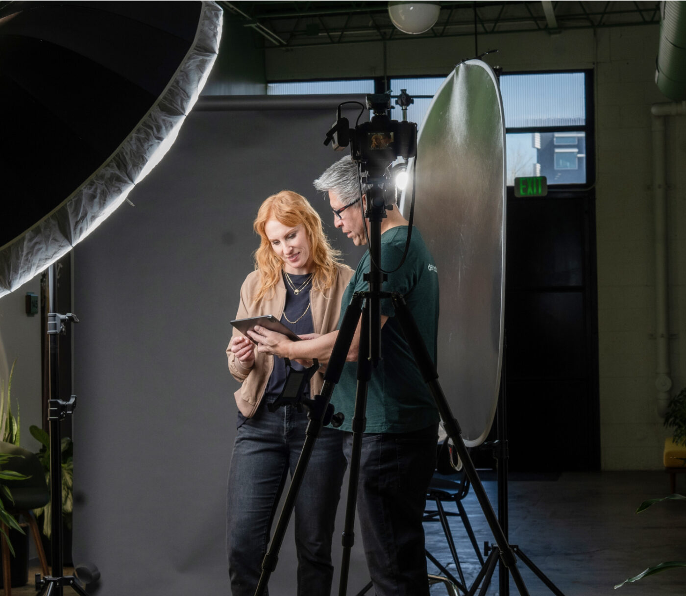 Two people from Denver Headshot Co are reviewing content on a tablet in a photography studio setup, complete with lights and a backdrop.