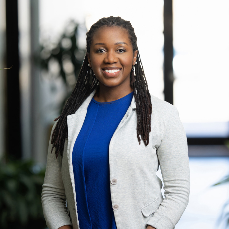 A woman with long braided hair, wearing a blue shirt and light gray blazer, stands smiling in a well-lit indoor setting, capturing the professional essence characteristic of Denver Headshot Co.