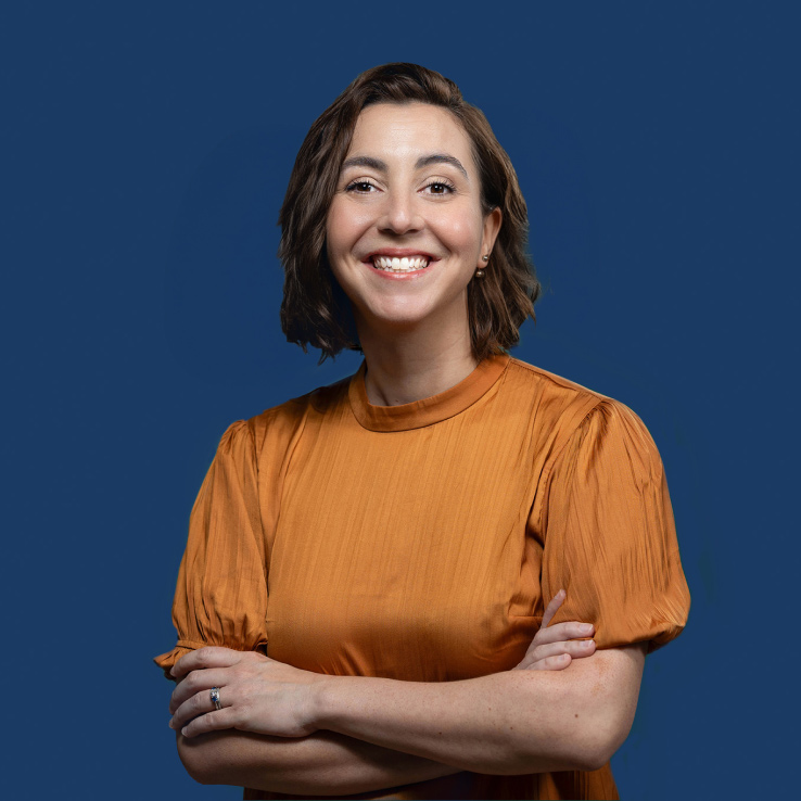 A woman with short brown hair smiles and crosses her arms. She is wearing an orange blouse and stands against a solid dark blue background, capturing the professional quality typical of Denver Headshot Co.