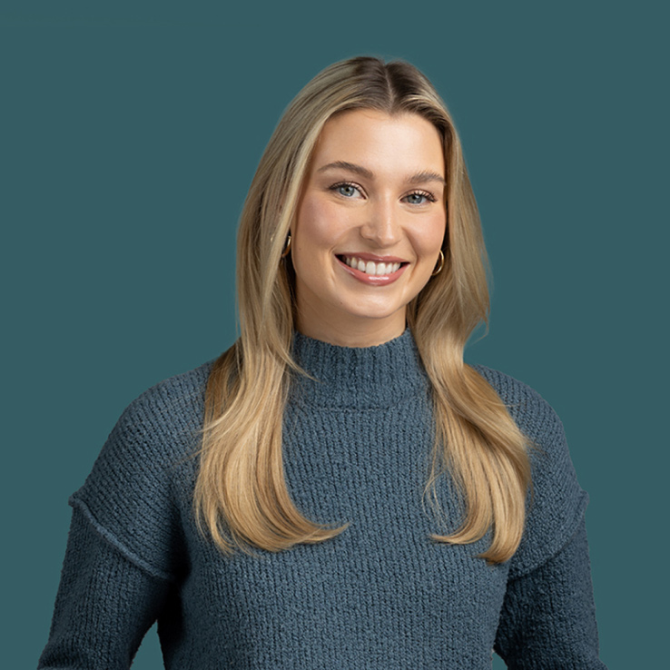 A woman with long blonde hair wearing a blue knit sweater smiles against a teal background, captured perfectly by Denver Headshot Co.