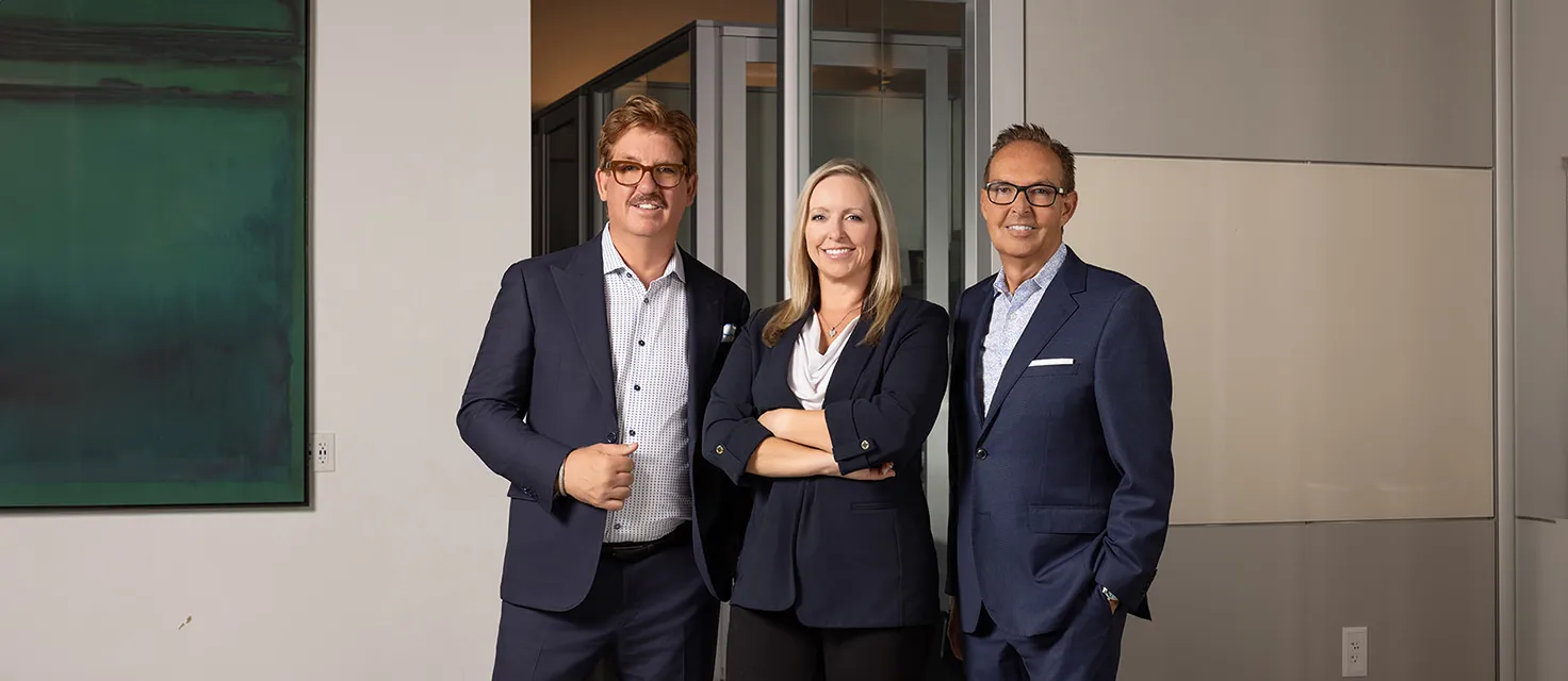 Three professionally dressed individuals, two men and one woman, stand together in an office setting, smiling at the camera.