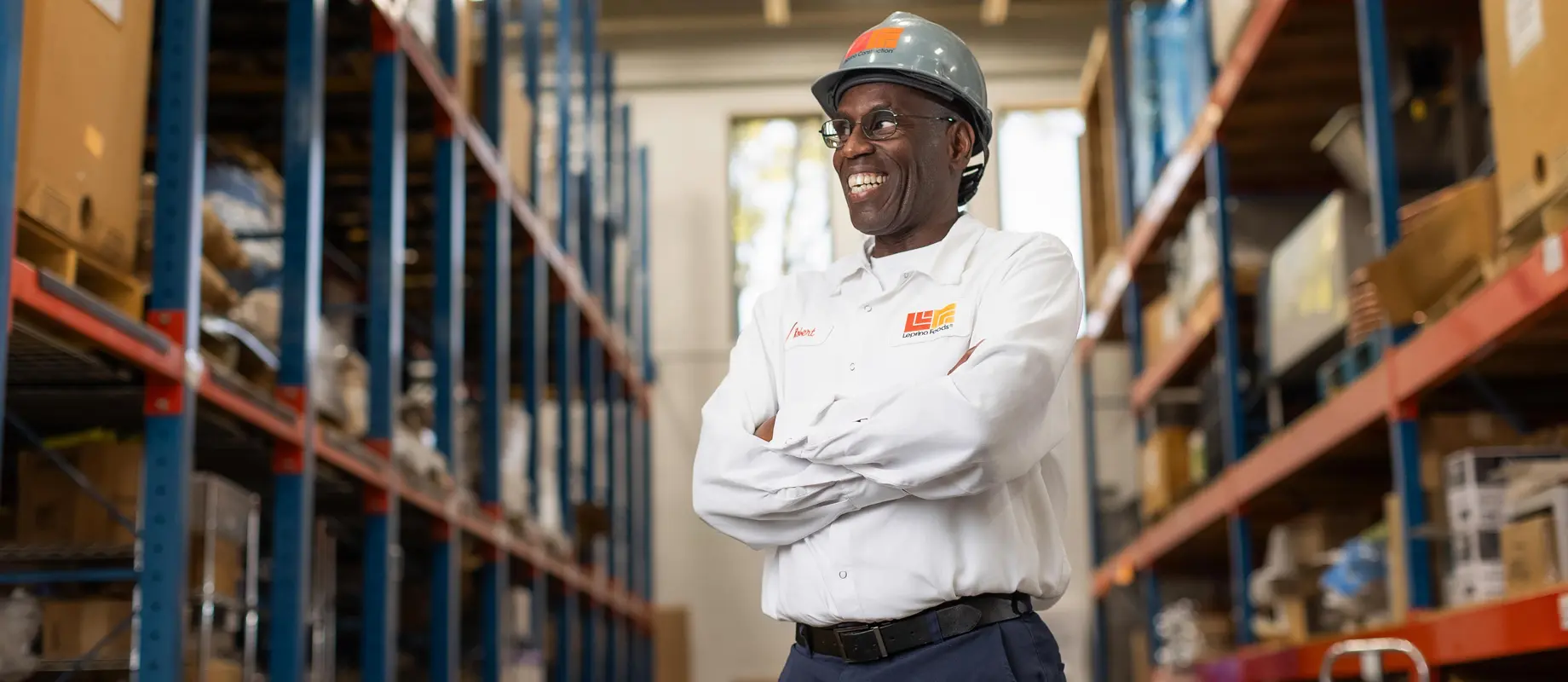 A man wearing a hard hat and safety glasses stands with arms crossed in a warehouse, smiling. He is dressed in a white company shirt and dark pants. Shelving with boxes lines the background.