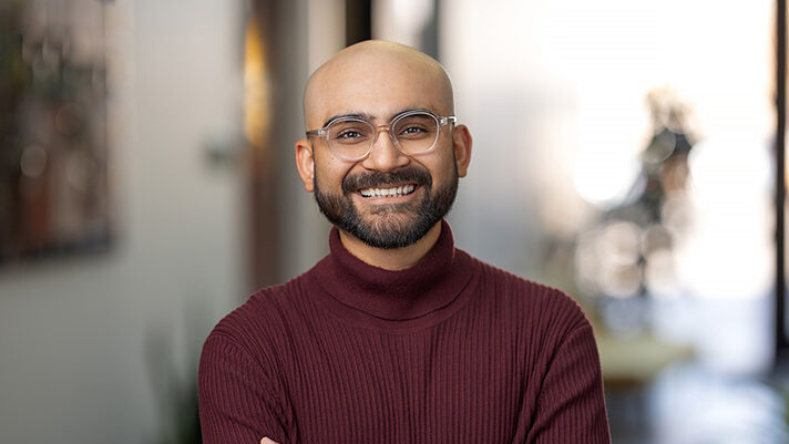 A person with glasses and a beard stands indoors, smiling with arms crossed, wearing a maroon sweater, perfectly captured by Denver Headshot Co.