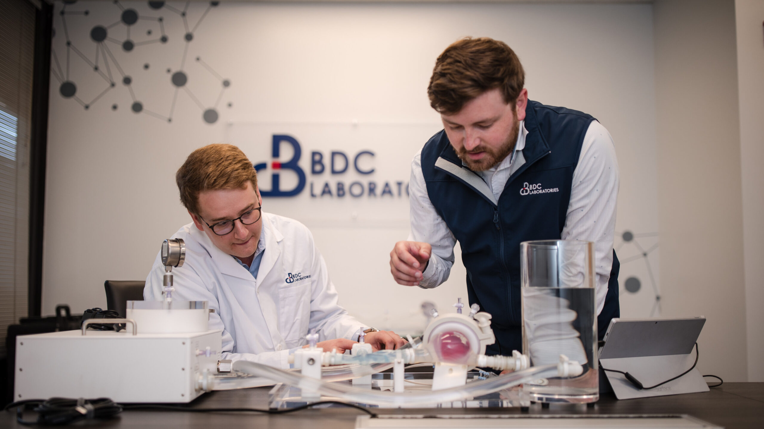 Two people in lab coats collaborate over scientific equipment at a table, with a video documenting their discoveries in the laboratory setting.